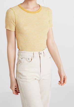 Krótki rękaw w żółto-białe paski z prążkowanym dekoltem, zestawiony z wysokim stanem jasnobeżowych dżinsów. Miękka tkanina o umiarkowanej elastyczności.