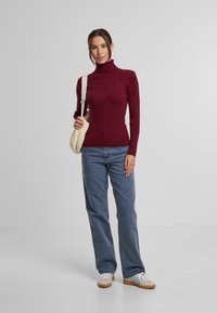 Vínová žebrovaná roláková mikina spárovaná s světle modrými volnými džíny a béžovými teniskami. Modelka drží krémovou crossbody kabelku.