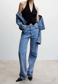 Schwarzes Halfter-Top, übergroße blaue Jeansjacke, hellblaue weite Jeans und schwarze spitze Schuhe vor einem schlichten Hintergrund.