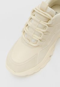 Beige chunky sneaker with thick sole, mesh tongue, and multiple lace loops, shown from a top-front angle on a white background.