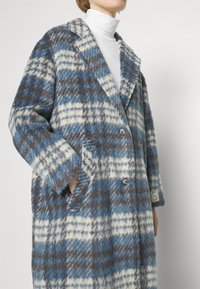 Kabát z vlněné směsi v modro-smetanovém kostkovaném vzoru, s klopovým límcem, dvěma knoflíky, velkými kapsami a měkkým texturovaným povrchem.