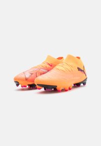 Scarpe da calcio di un brillante arancione, con una parte superiore testurizzata, colletto simile a una calza e suola nera con accenti rosa e tacchetti per la trazione.