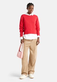 Mujer vistiendo un suéter rojo brillante sobre una camisa blanca, pantalones de cortocultura beige, zapatillas blancas y sosteniendo un bolso rosa claro contra un fondo liso.