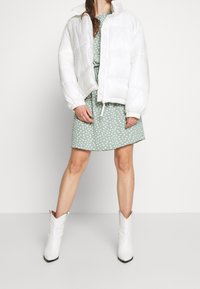 Femme portant une doudoune blanche sur une robe verte à pois blancs, associée à des bottines blanches à bout pointu, se tenant devant un fond uni.