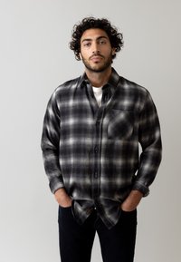 Zwart-wit geblokt flanellen overhemd met een borstzak, knoopsluiting en opgerolde mouwen, gedragen over een wit T-shirt.