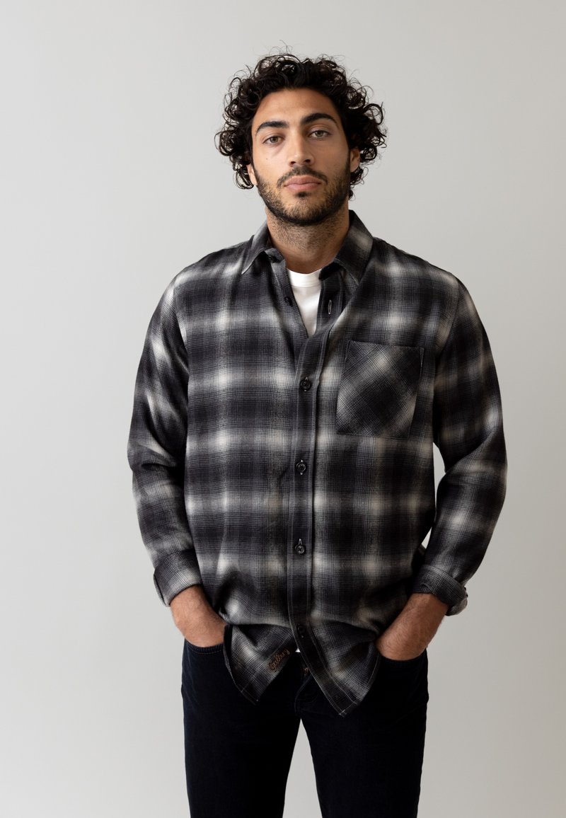Zwart-wit geblokt flanellen overhemd met een borstzak, knoopsluiting en opgerolde mouwen, gedragen over een wit T-shirt.