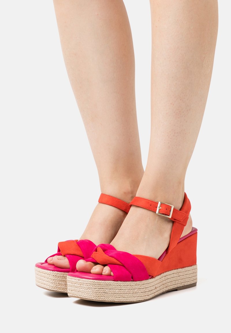 Pieds portant des sandales compensées rouges et roses avec des brides à la cheville et des semelles en jute tressé sur un fond clair.