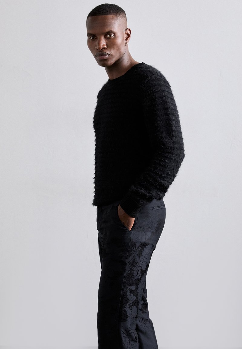 Sort strikket sweater med teksturerede striber, parret med sorte bukser med et diskret blomstermønster og en tætsiddende pasform.