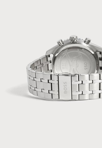 Montre bracelet en métal argenté avec logo BOSS sur la boucle, montrant le verso du cadran avec un motif gravé et des boutons.