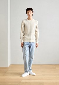 Beżowy sweter w splot warkoczowy z długimi rękawami, w zestawieniu z jasnoniebieskimi dżinsami i białymi trampkami. Prosty design i widoczna faktura materiału.