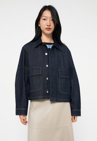 Ung kvinne med rett svart hår har på seg en mørkeblå denimjakke med knapper og beige skjørt, vendt mot kameraet foran en ensfarget hvit bakgrunn.