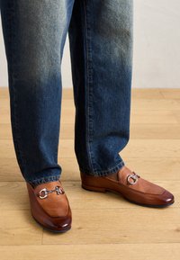 Pantofi loafers din piele maro cu vârful strălucitor și detalii din argint, asortați cu blugi de denim de culoare bleu deschis, pe un podea din lemn.