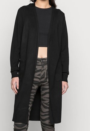 Vrouw draagt een lange zwarte open cardigan, een zwart cropped top en grijs-zwarte zebraprint broek tegen een effen achtergrond.