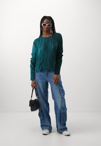 Top lungo a maniche lunghe plissettato in teal abbinato a jeans cargo azzurri chiari, sneakers bianche e una piccola borsa nera. Presenta trecce spesse e occhiali da sole bianchi.