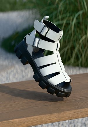 Weiße Sandale mit verstellbaren Riemchen, robuster schwarzer Gummisohle und Peeptoe-Design auf einer Holzoberfläche.