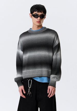 Młody mężczyzna w czarnym swetrze z gradientem, niebieskiej podkoszulce, czarnych spodniach z kluczami na karabińczyku i czarnych okularach przeciwsłonecznych na jednolitym tle.