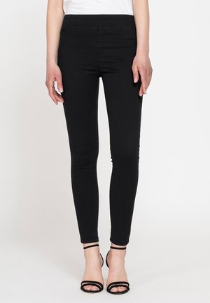 Donna che indossa pantaloni neri aderenti e sandali neri con tacco alto e cinturini, in piedi su sfondo bianco.