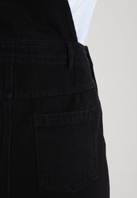 Gros plan sur la poche et la taille d'une salopette en denim noir portée sur une chemise bleu clair sur un fond uni.