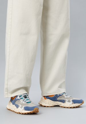 Persona che indossa pantaloni bianchi a gamba larga e sneakers blu, grigie e arancioni con suole bianche testurizzate, in piedi su pavimento grigio.