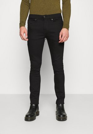 Pantalones vaqueros negros ajustados fabricados en denim, con un diseño clásico de cinco bolsillos, cierre con botón y un ligero estiramiento para mayor comodidad. Se llevan con botas negras.