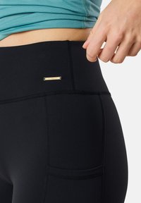 Des leggings noirs fabriqués en tissu extensible présentent une taille haute, des coutures plates et un petit logo doré en accent. Un haut texturé est visible au-dessus.