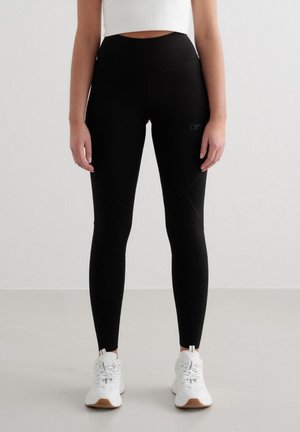 Kvinne iført svarte høytlivs leggings og hvite joggesko, stående mot en ensfarget lys grå vegg og gulv.