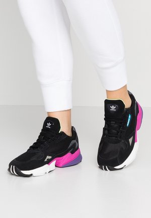 Czarne buty sportowe Adidas z różowo-fioletowymi podeszwami, noszone z białymi spodniami na jasnym tle, z widocznym logo Adidas na językach.