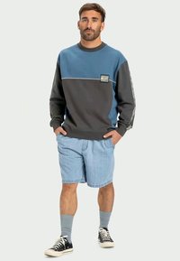 Bluza w niebiesko-szarym kolorze z okrągłym dekoltem, kontrastowym panelem i naszywką z logo. W zestawie z jasnymi szortami dżinsowymi i czarnymi trampkami.
