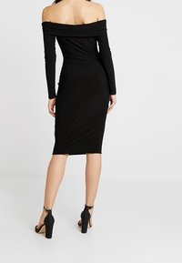 Žena v upnutých černých midi šatech s dlouhým rukávem a bez ramínek, doplněná černými lodičkami na vysokém podpatku se zapínáním kolem kotníku, stojící zády.