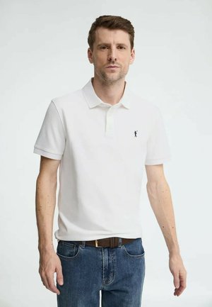 Man met kort bruin haar, gekleed in een wit poloshirt met een klein zwart logo op de borst en een blauwe spijkerbroek, staand tegen een effen lichte achtergrond.