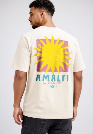 Mann trägt cremefarbenes T-Shirt mit großer gelber Sonne und dem Schriftzug „AMALFI La Dolce Vita“ auf dem Rücken, kombiniert mit dunklen Jeans.