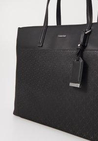 Sac fourre-tout noir en similicuir lisse avec un motif embossé. Il est équipé de poignées solides et d'une plaque logo argentée pour l'identification de la marque.