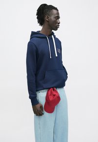 Άνδρας με ντραντλόκς που φοράει μια ναυτική μπλε φούτερ με κουκούλα και ανοιχτόχρωμο τζιν, κρατώντας κόκκινο καπέλο στο πλάι του, στραμμένος προς τα δεξιά μπροστά σε απλό λευκό φόντο.