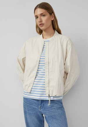 Kobieta ubrana w kremową kurtkę na zamek, na którą nałożona jest jasnoniebieska i biała koszula w paski oraz niebieskie dżinsy, stojąca na jednolitym szarym tle.