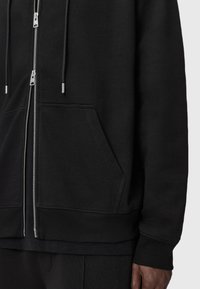 Haut à capuche noir zippé en tissu doux ; comprend une poche kangourou, un ourlet côtelé et des détails de fermeture éclair en métal.