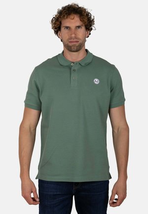Hombre de pelo rizado lleva un polo verde de manga corta con un pequeño logo redondo en el pecho, combinado con vaqueros oscuros y fondo neutro.