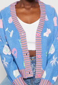 Blå strikket cardigan med legende print såsom muslinger og palmer. Stripet krave og ærmer i pink og grøn, med pink knapper.