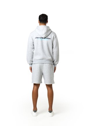 Mann mit dem Rücken zur Kamera, trägt einen hellgrauen Kapuzenpullover mit dem Schriftzug "closurelondon" und passende Shorts, weiße Sneaker auf weißem Hintergrund.