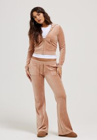 Twee-delig loungewear-set in lichtbeige, bestaande uit een korte rits hoodie en uitlopende broeken met voorvakjes, gemaakt van zachte fluwelen stof.