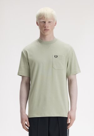 Jeune personne à la peau pâle et aux cheveux platine portant un t-shirt à manches courtes vert clair avec une poche poitrine et un pantalon foncé.