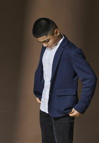 Blazer marine avec des épaules structurées, des revers crantés et deux poches avant. Porté sur une chemise blanche à boutons et un jean foncé.