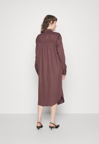 Vestido longo, cor bordô, com costas ajustadas, colarinho abotoado, mangas longas e uma bainha assimétrica. Tecido liso com detalhes mínimos.