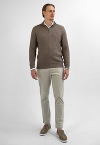 Bruine kabelgebreide pullover, kwartrits, lange mouwen, slim-fit beige broek en beige instappers. Witte kraag is zichtbaar.