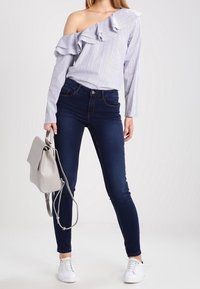 Top a righe senza spalle con volant, jeans attillati blu navy e sneakers bianche. Una borsa grigia pende dalla spalla sinistra.