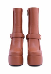 Stivali alla caviglia in finta pelle marrone con una texture liscia, punta quadrata, suola a piattaforma rialzata e cerniere laterali. Presentano un'ampia fascia decorativa.
