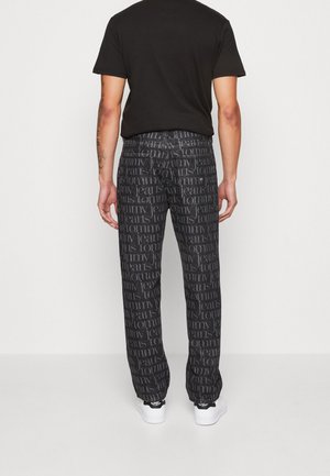 Mand stående med ryggen til, iført sorte trykte jeans med teksten "Tommy Jeans", sort t-shirt og hvide sneakers.