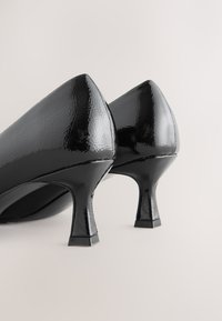 Crne salonke od lakirane kože s oštrim vrhom i debelom potpeticom. Cipele imaju glatki, sjajni završetak i klasični siluetu.