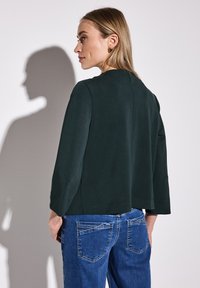 Temně zelený, lehký top s kulatým výstřihem, tříčtvrtečními rukávy a volným střihem, kombinovaný s modrými džíny na středním pasu.
