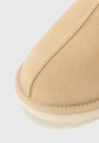 Details van een beige suède schoen met tonale stiksels, een ronde neus en een lichte rubberen zool. De textuur is zacht en glad.