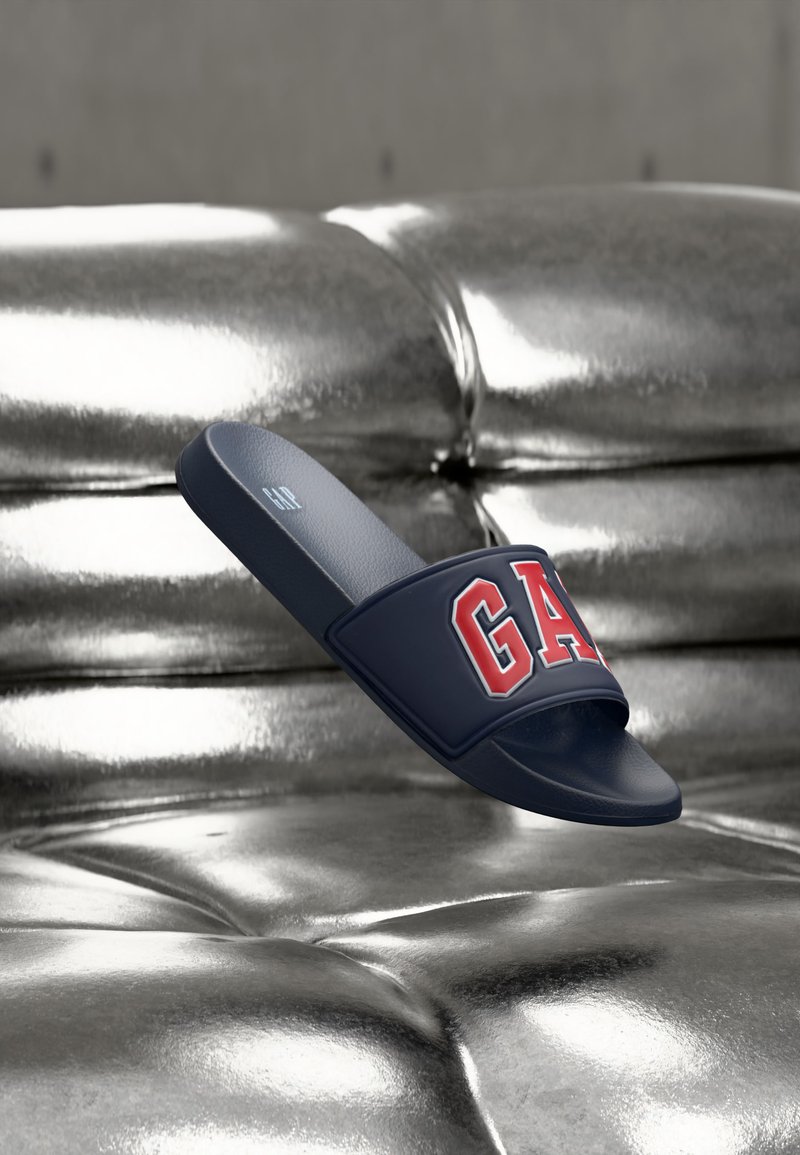 Sandale à glissière bleu marine avec une bride rouge portant le logo "GAP", flottant au-dessus d'une surface en cuir argenté capitonné réfléchissante.